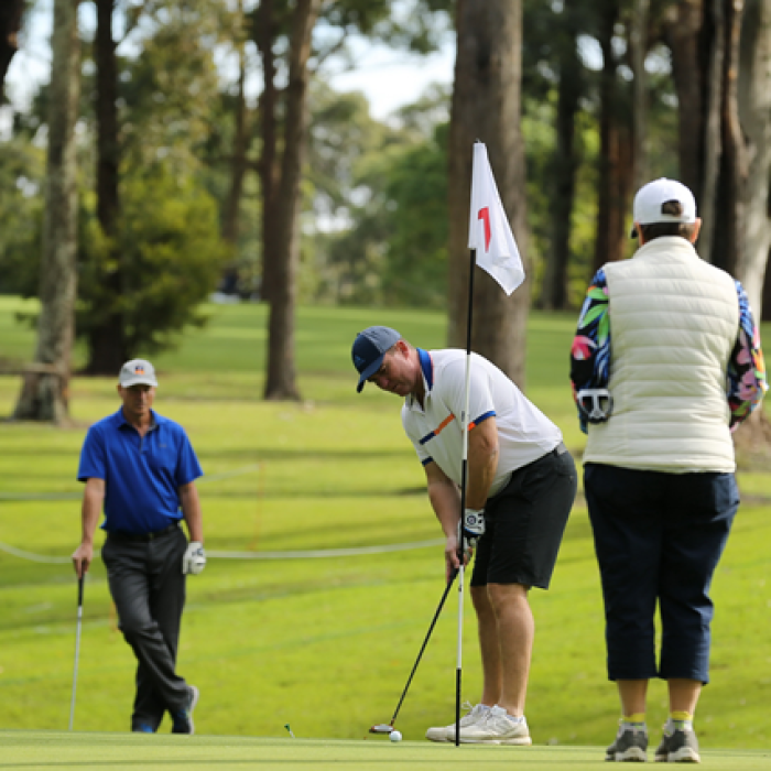 Three people on a golf course, one putting near a flag while others watch.
